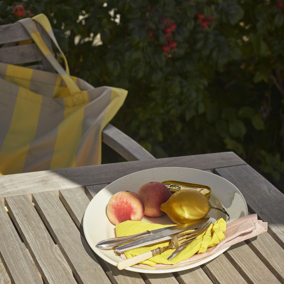 Yellow striped tote bag, pastel fruits on a cream plate, and pink utensils on a wooden table under soft sunlight.