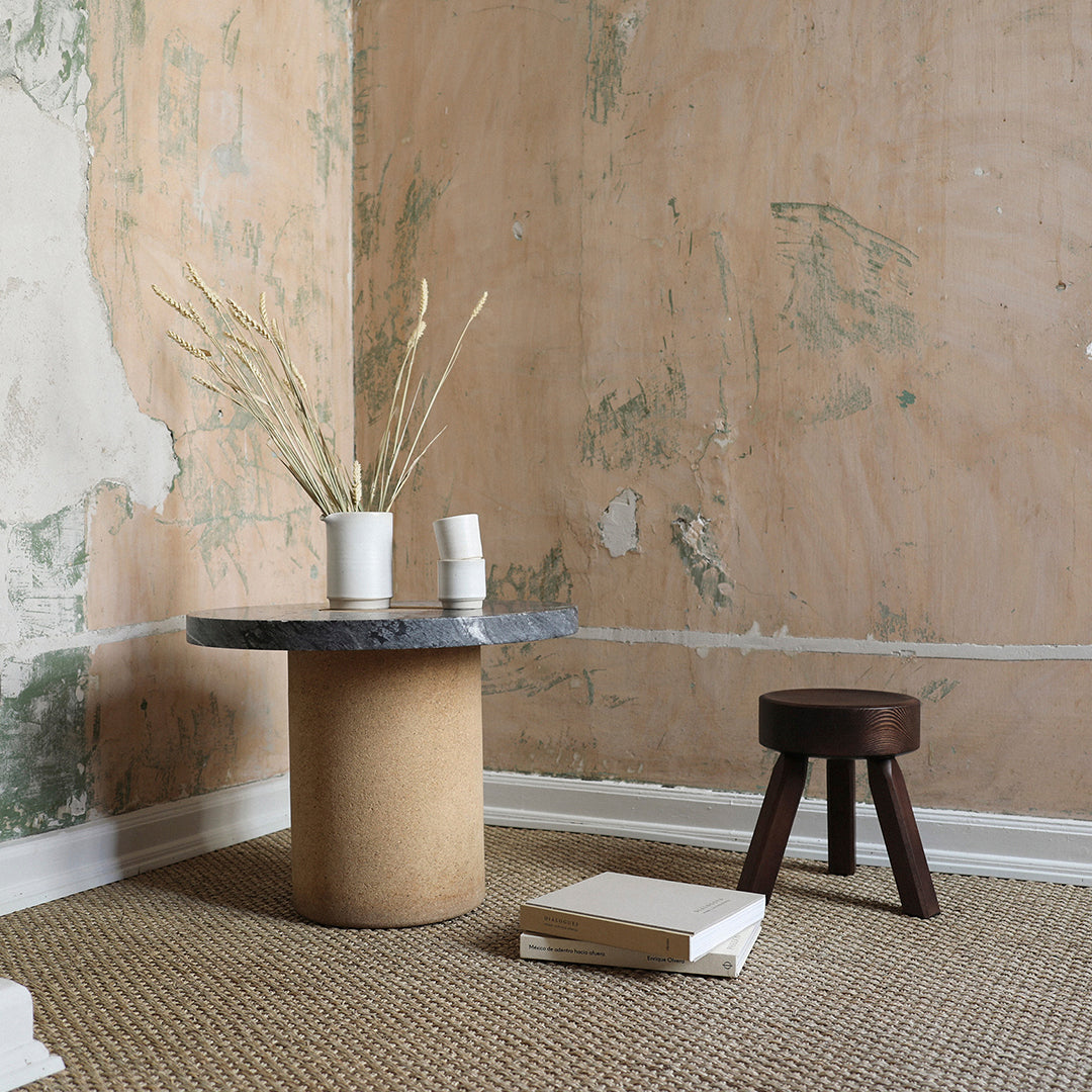Circular gray marble tabletop on a textured beige base, complemented by a dark brown stool in a minimalist setting.
