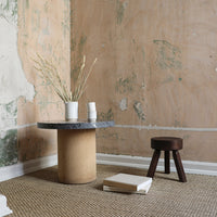 Circular gray marble tabletop on a textured beige base, complemented by a dark brown stool in a minimalist setting.