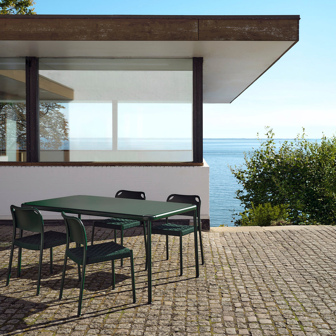 Green outdoor dining table and chairs set against a sunny coastal backdrop with a modern architectural design.