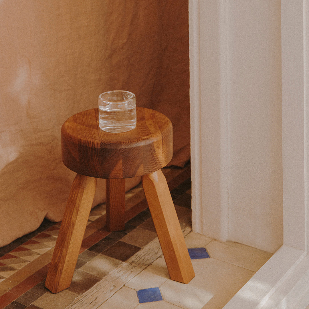 Wooden stool with a round top and light brown legs, featuring textured wood grains and a glass sitting on top.