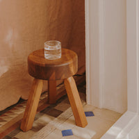 Wooden stool with a round top and light brown legs, featuring textured wood grains and a glass sitting on top.