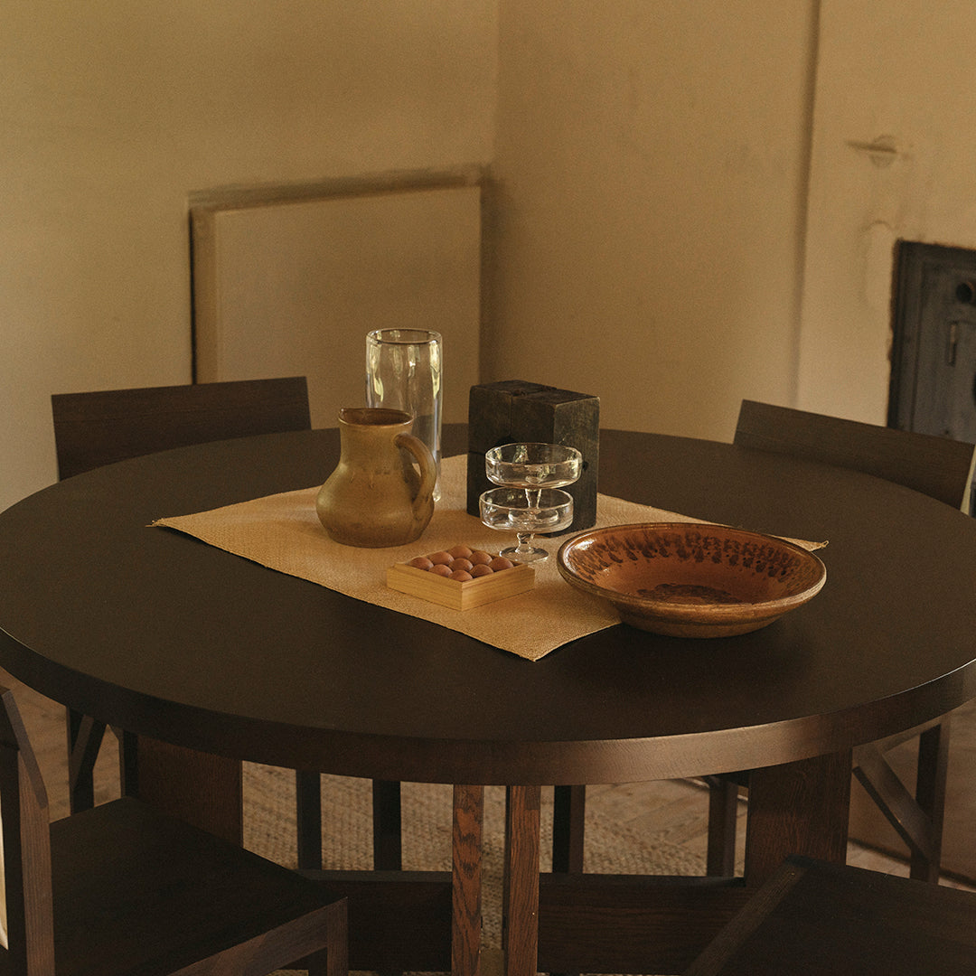 Round dark wooden dining table with earthy-toned ceramics and clear glassware, featuring a beige table runner.