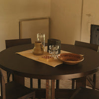 Round dark wooden dining table with earthy-toned ceramics and clear glassware, featuring a beige table runner.