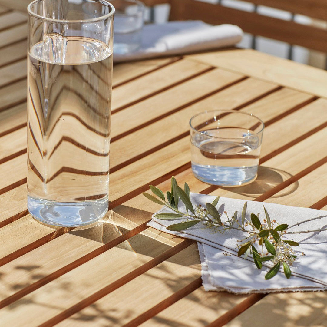 Clear glassware atop a textured wooden table with parallel light and shadow patterns, accented by green olive leaves.