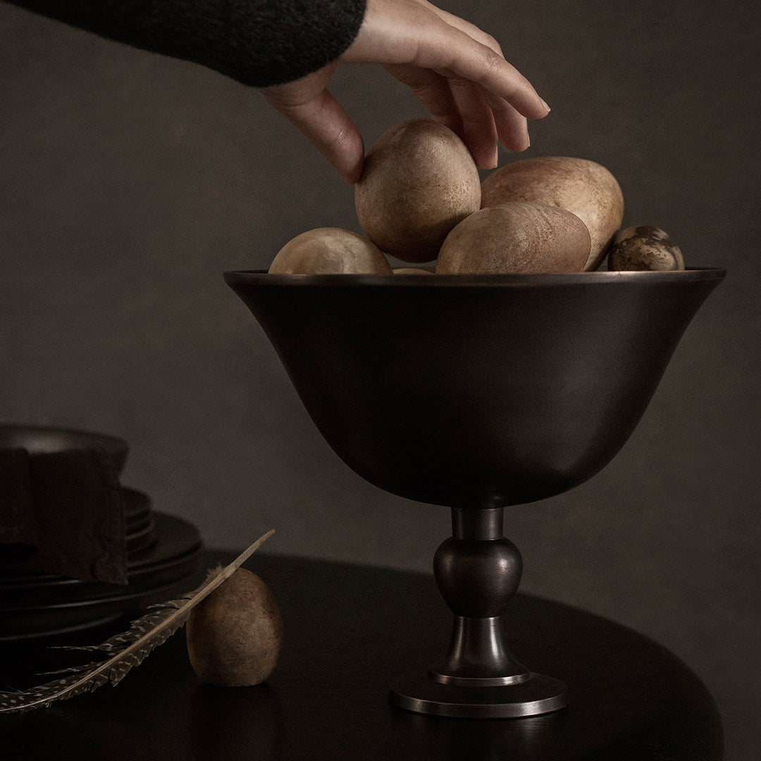 "Dark matte bowl filled with textured earth-toned stones, set against a muted backdrop and elegant tableware."