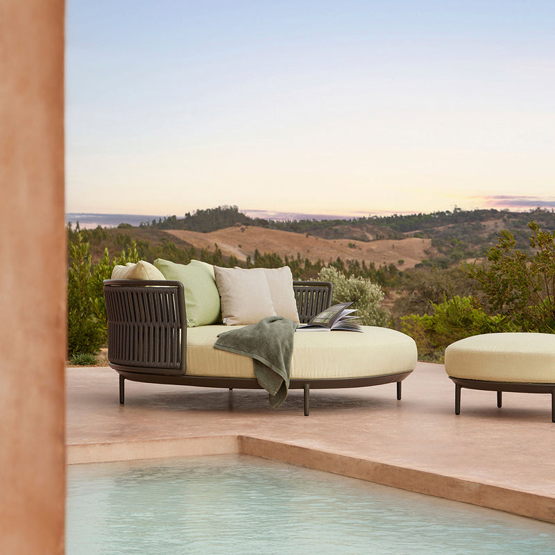 "Round outdoor sofa in light green with neutral cushions and dark woven detailing, complemented by serene natural scenery."