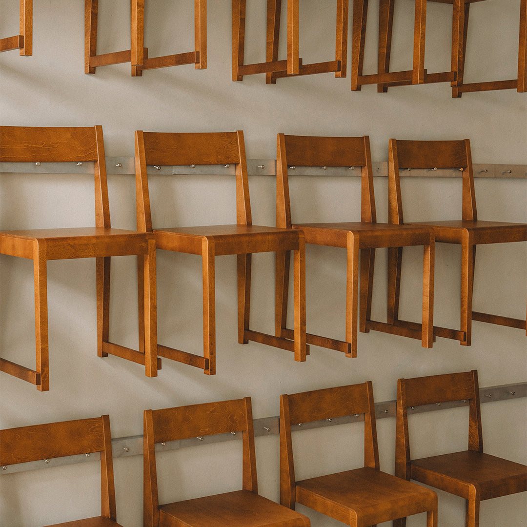 Wooden chairs in a warm brown finish, displayed in a minimalistic, stacked arrangement against a neutral wall.