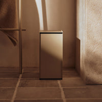 Sleek gold trash can with a smooth finish, set against warm, neutral-toned surroundings and subtle textures.