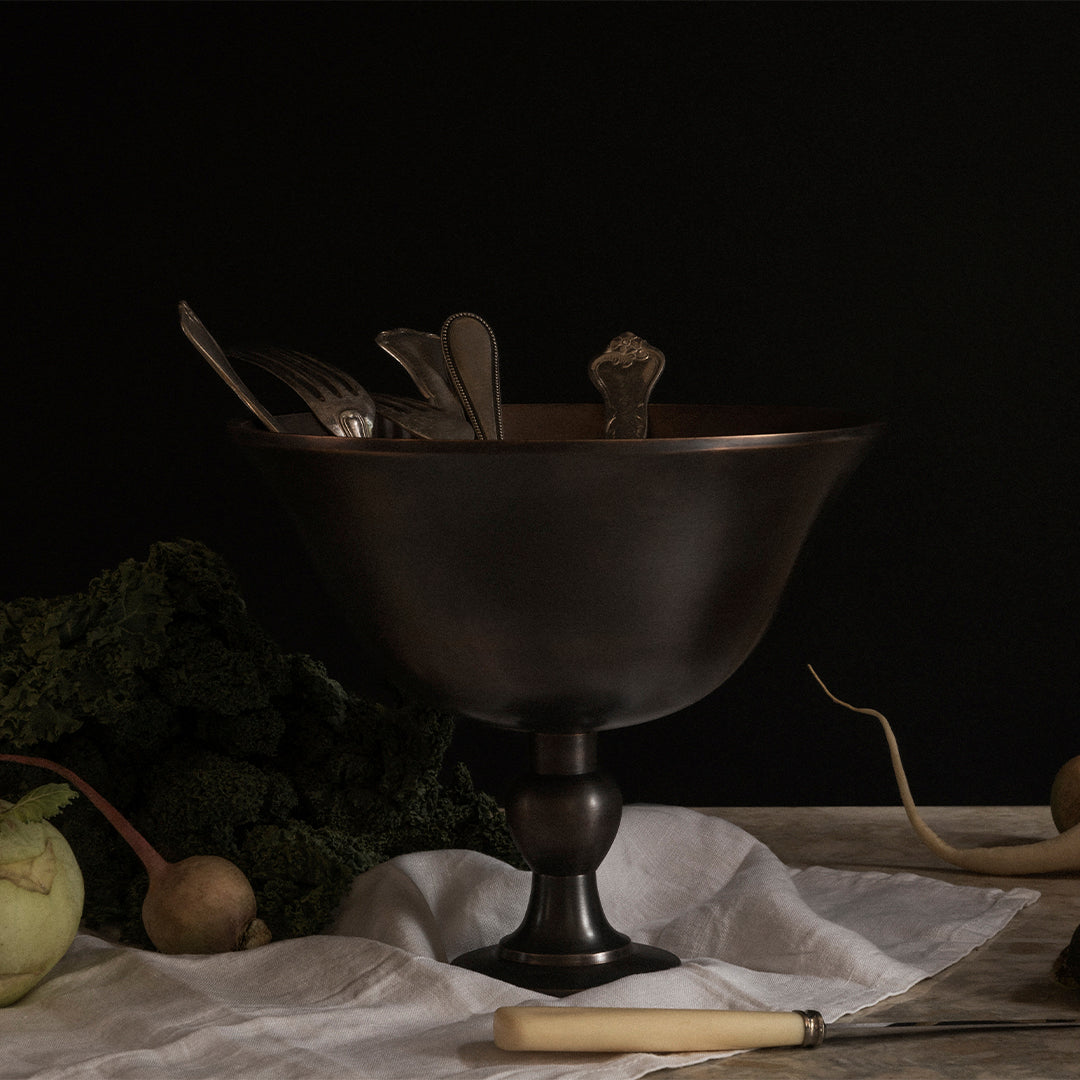 Black and bronze bowl on a dark background, complemented by silver utensils and earthy greens. Elegant vintage design.