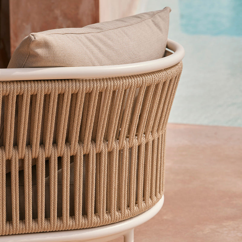 Beige woven rope chair with a cream cushion against a light pink surface and turquoise water backdrop.