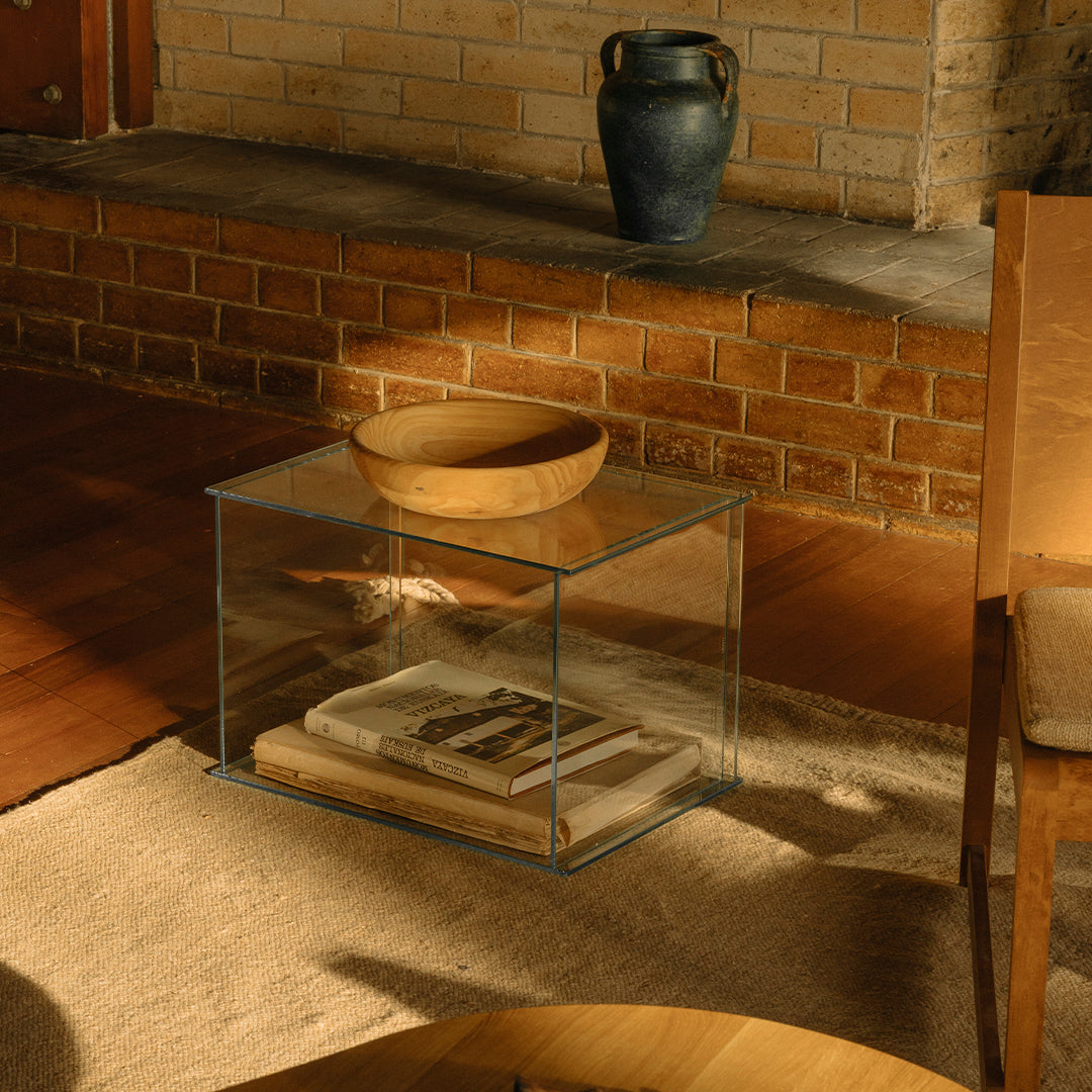 Clear glass coffee table paired with a round wood bowl, set against earthy brick and warm wood textures.