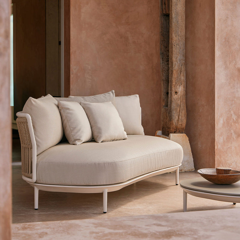 Beige curved sofa with three matching cushions on a light-toned floor and earthy wall backdrop. Modern and minimalist design.