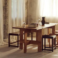 Wooden dining table with natural finish, complemented by black and wooden stools against soft beige and white drapery.