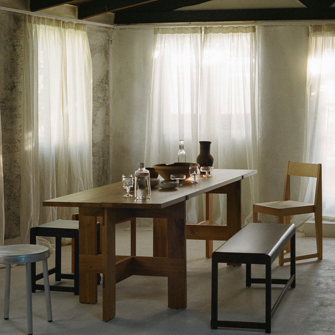 Wooden dining table in warm tones, paired with neutral chairs and benches, set against soft, light-filtering curtains.