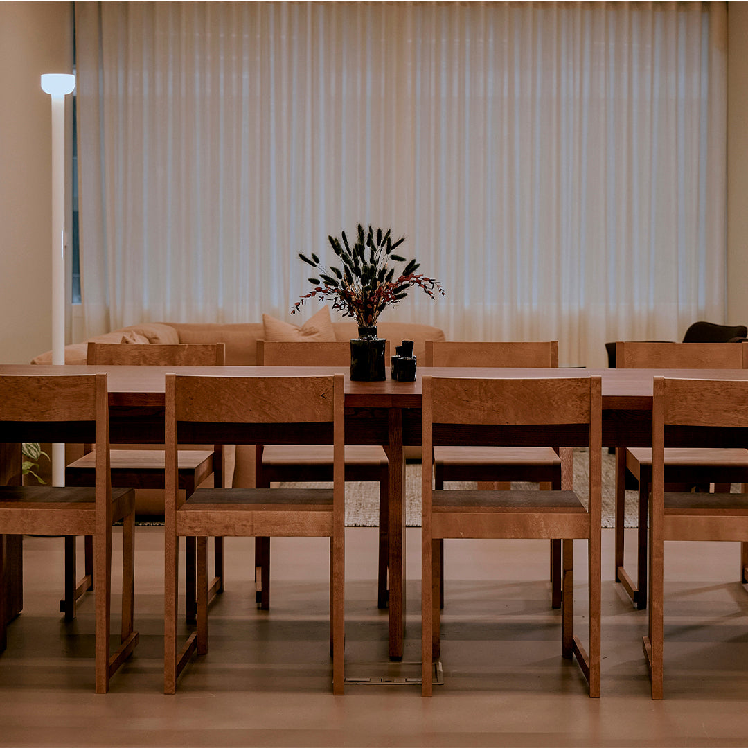 Wooden dining table set with neutral-toned fabric chairs, complemented by a simple black vase and muted curtains.