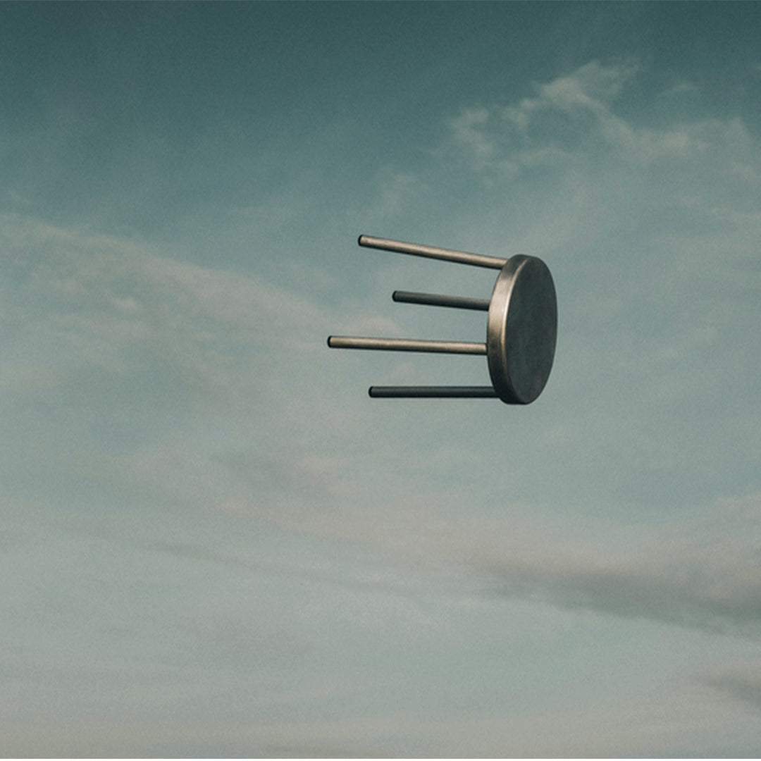 Floating gray stool with a smooth, circular top and three metallic legs against a soft blue sky.