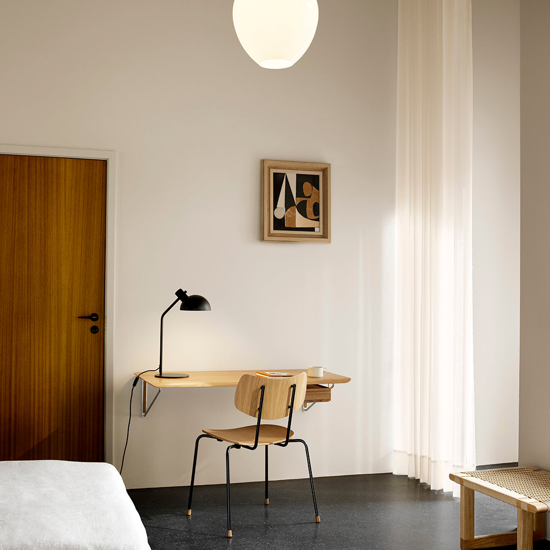 Minimalist interior featuring neutral walls, light wood tones, and a structured black desk lamp against a bright backdrop.