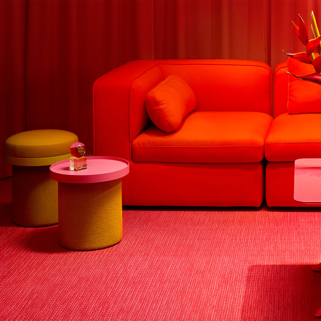 Bright red couch with orange cushions, yellow round stool, and pink accent table against a red backdrop and textured pink rug.