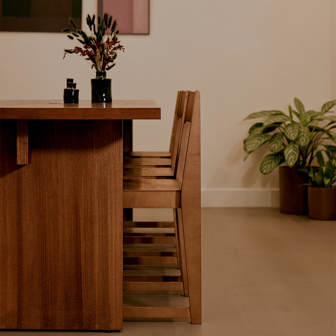 Wooden dining table with natural finish, paired with beige chairs and green plants, in a warm, minimalist setting.