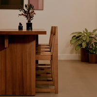 Wooden dining table with natural finish, paired with beige chairs and green plants, in a warm, minimalist setting.