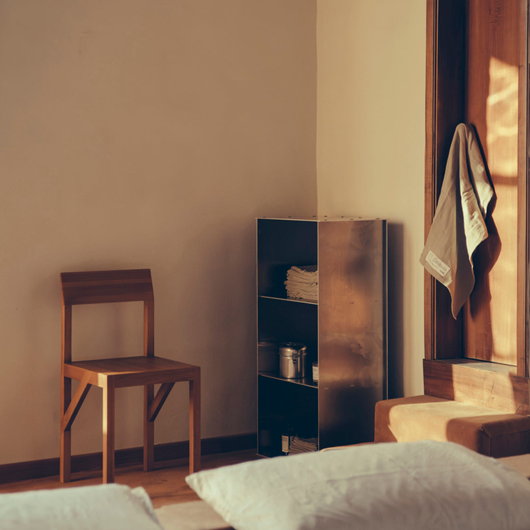 Warm beige walls, light wooden chair, sleek black bookshelf, soft greys in bedding, and a white towel hanging.
