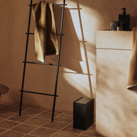 "Minimalist bathroom scene with earthy tones, black ladder, and beige textured walls casting soft shadows."