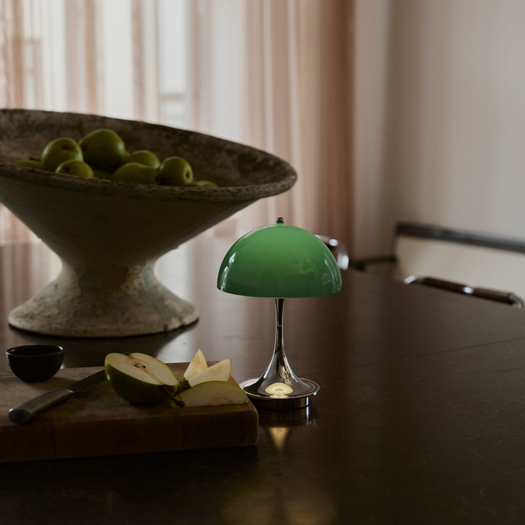 Green glass lamp with a shiny chrome base, set against a neutral backdrop featuring a textured bowl of green apples.