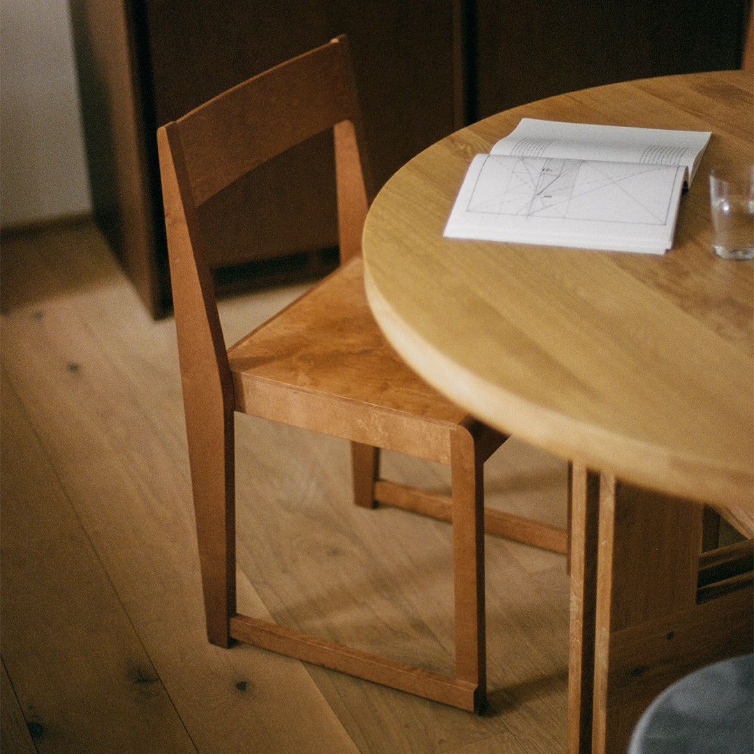Modern wooden chair in warm oak tones next to a round light wood table, complemented by natural flooring textures.