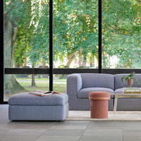 Light blue plaid sofa with matching ottoman and a coral round stool on a neutral rug, surrounded by lush greenery.