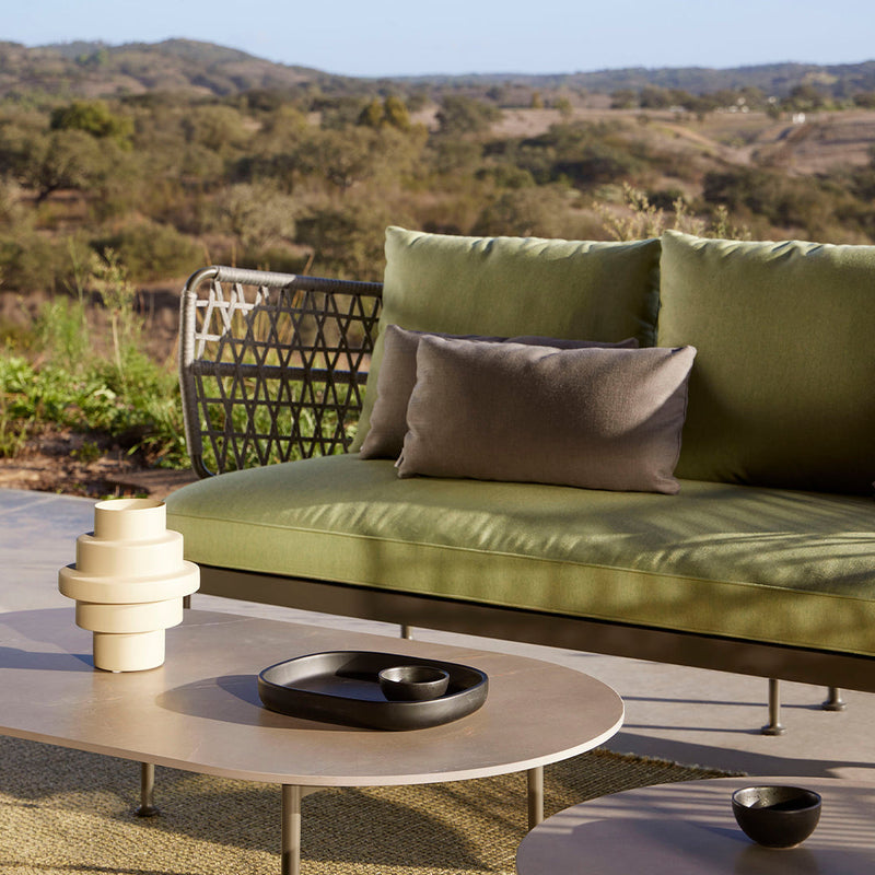 Green outdoor sofa with neutral cushions, layered textures, and sleek, modern table accents. Natural landscape in background.