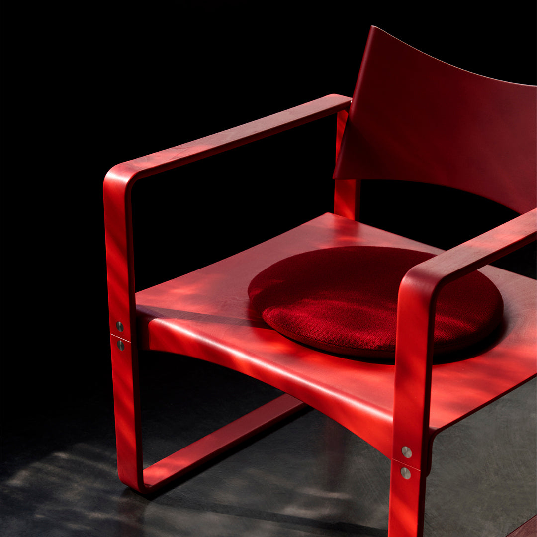 Red wooden chair with a round red cushion, featuring sleek lines and a minimalist design against a dark background.