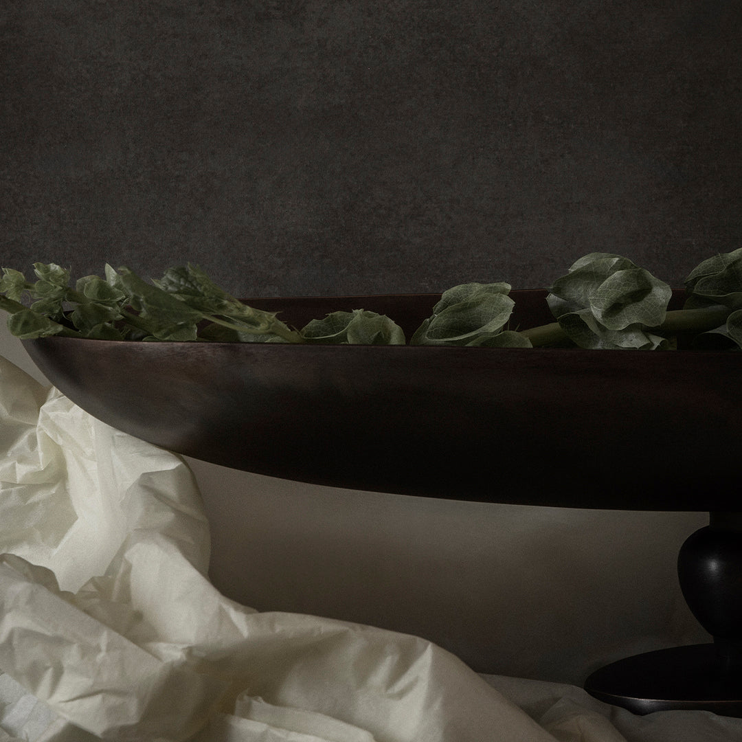 Dark brown decorative bowl with green leaves, set against a textured gray background and soft white fabric.