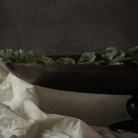 Dark brown decorative bowl with green leaves, set against a textured gray background and soft white fabric.