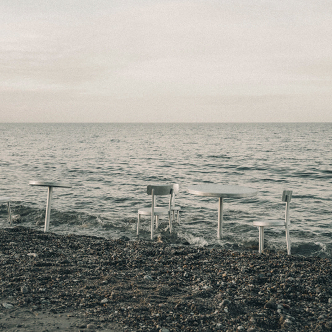 Minimalist outdoor table set in soft gray tones with a serene water backdrop and subtle textured ground.