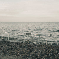 Minimalist outdoor table set in soft gray tones with a serene water backdrop and subtle textured ground.