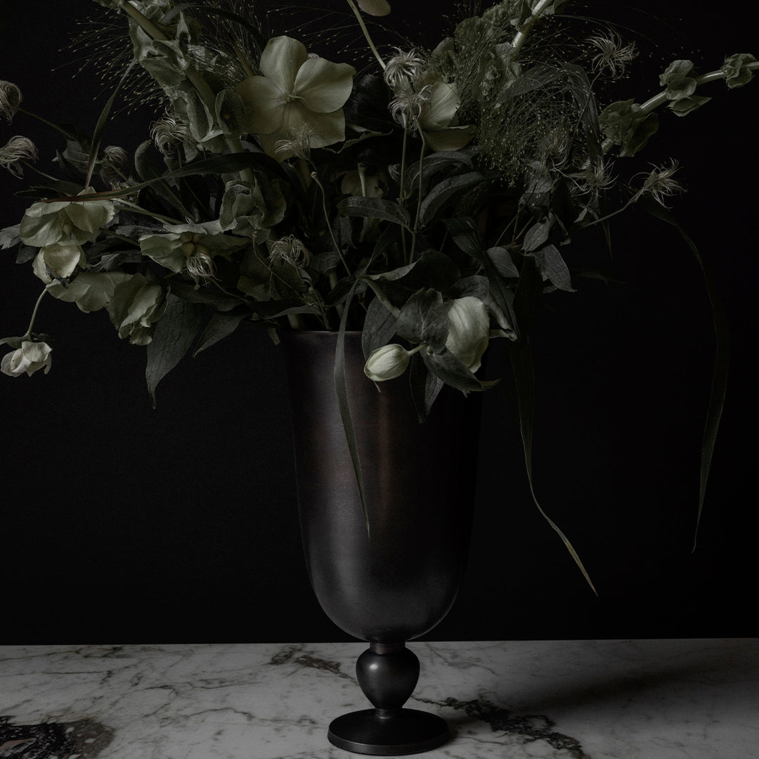"Dark floral arrangement in a sleek black vase, featuring soft greens and creamy white blossoms against a black background."