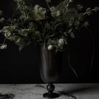 "Dark floral arrangement in a sleek black vase, featuring soft greens and creamy white blossoms against a black background."