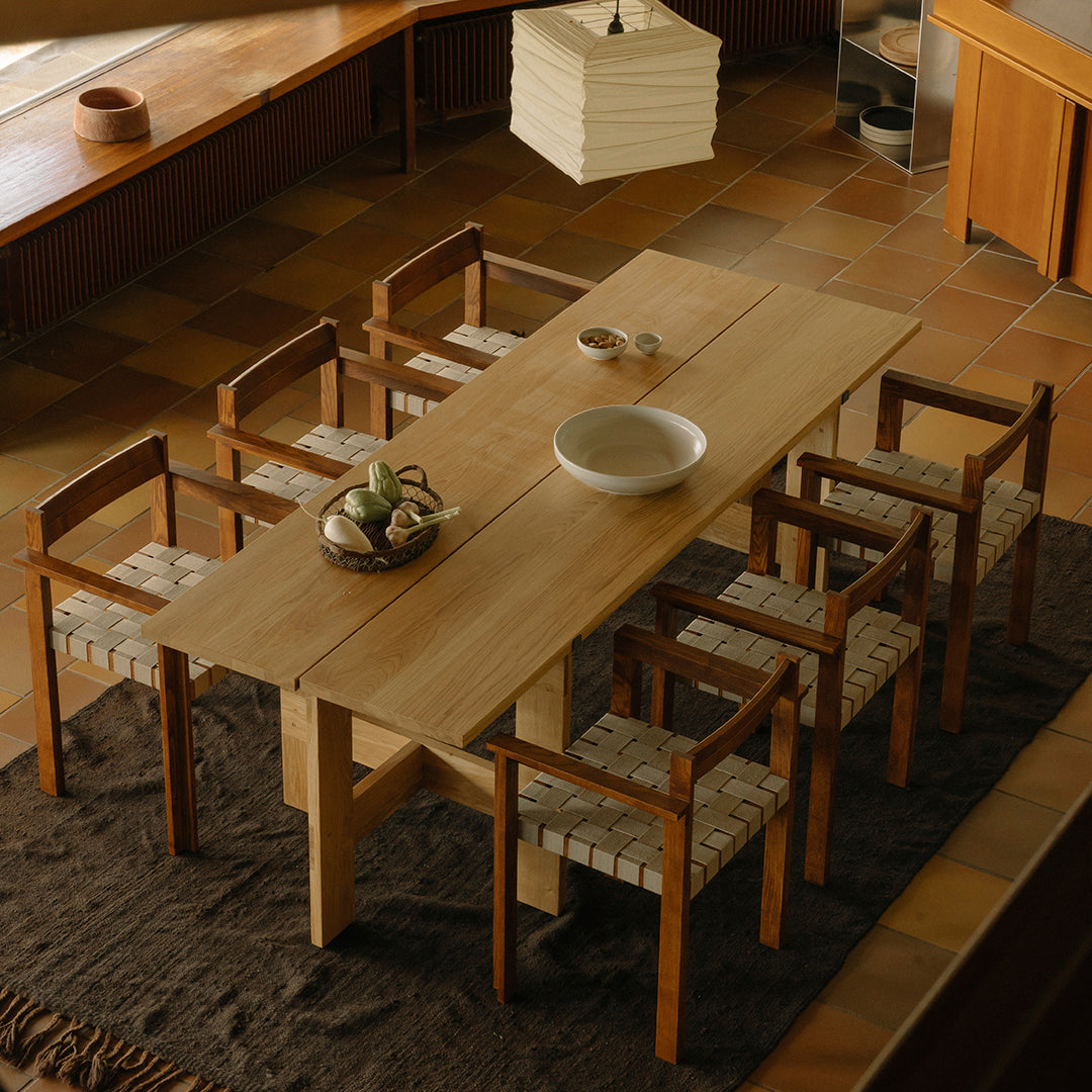 Natural wood dining table with beige woven chairs on a textured brown rug, set in a warm, earthy-toned room.