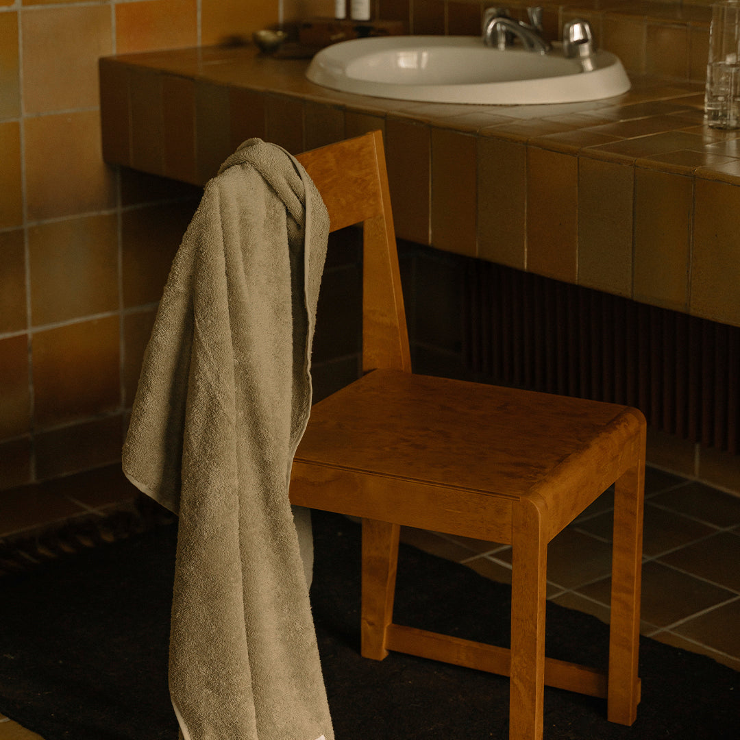 Beige towel draped on a warm wooden chair in a neutral-toned bathroom with tiled surfaces and rustic patterns.
