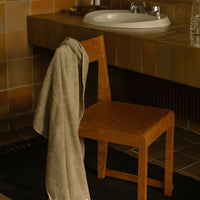 Beige towel draped on a warm wooden chair in a neutral-toned bathroom with tiled surfaces and rustic patterns.