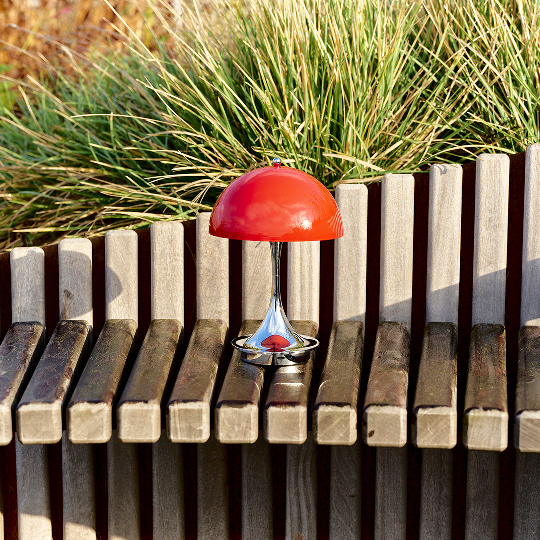 Red mushroom-shaped lamp with a shiny chrome base, set against a backdrop of wooden slats and green grass.