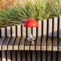 Red mushroom-shaped lamp with a shiny chrome base, set against a backdrop of wooden slats and green grass.