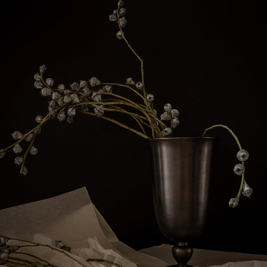 Dark metal vase with delicate gray floral stems against a black background, featuring subtle textures and elegant shapes.
