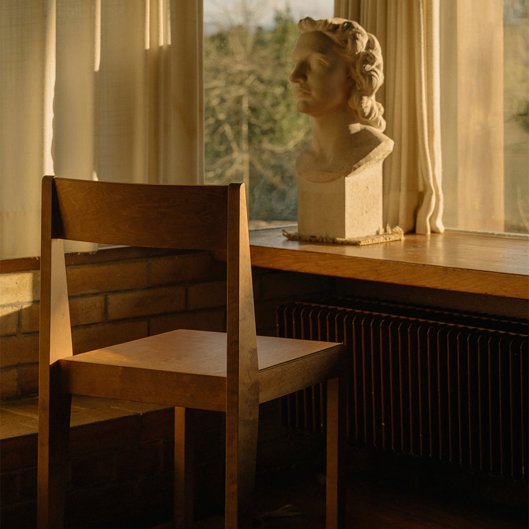 Wooden chair in warm tones beside a sunlit window, featuring a simple, minimalist design.