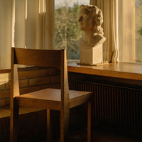 Wooden chair in warm tones beside a sunlit window, featuring a simple, minimalist design.