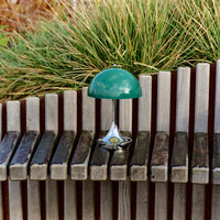 Green dome lamp with a sleek chrome base, set against wooden slats and lush greenery. Modern design and vibrant colors.