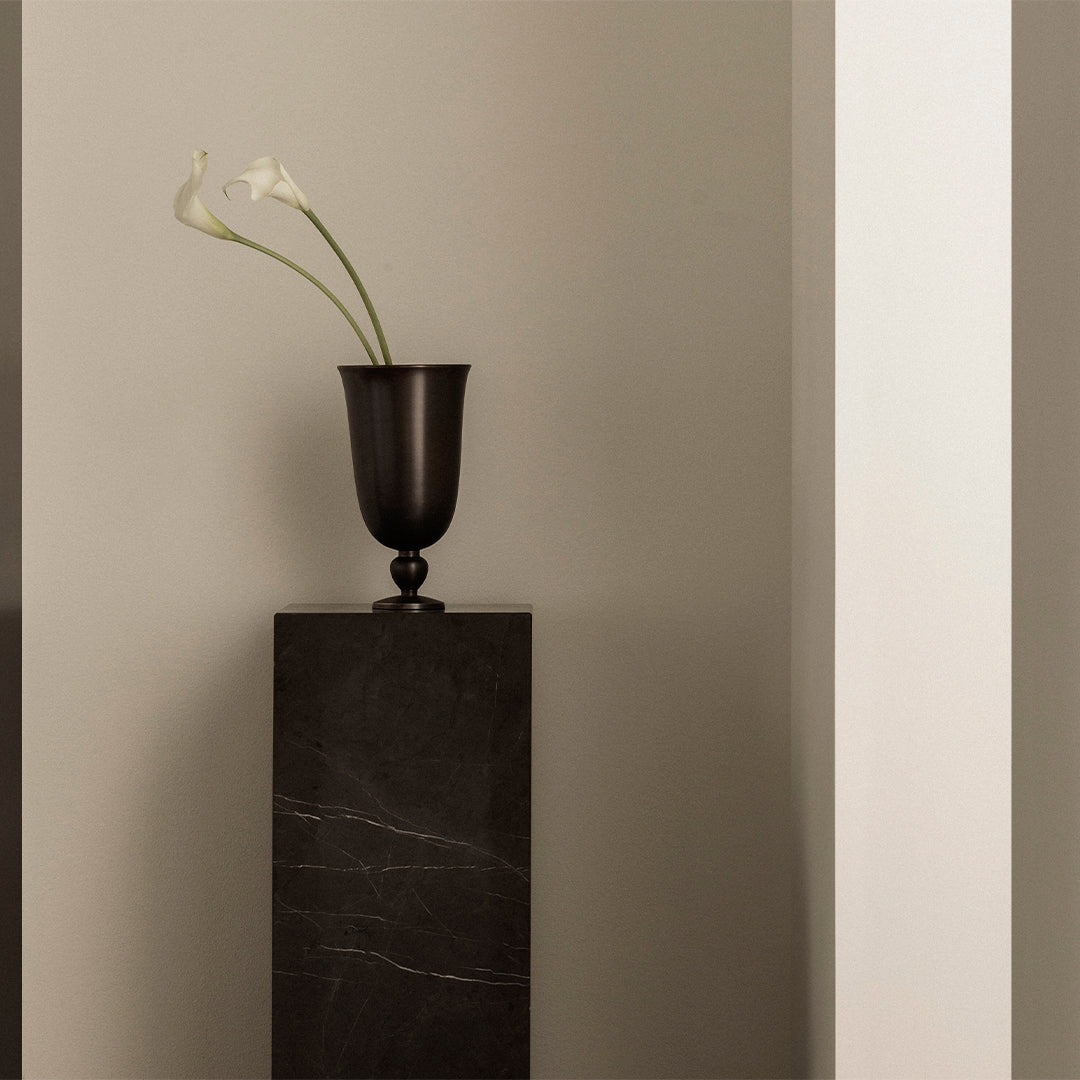Black vase with white flowers on a dark stone pedestal against a neutral gray wall, showcasing elegant minimalism.