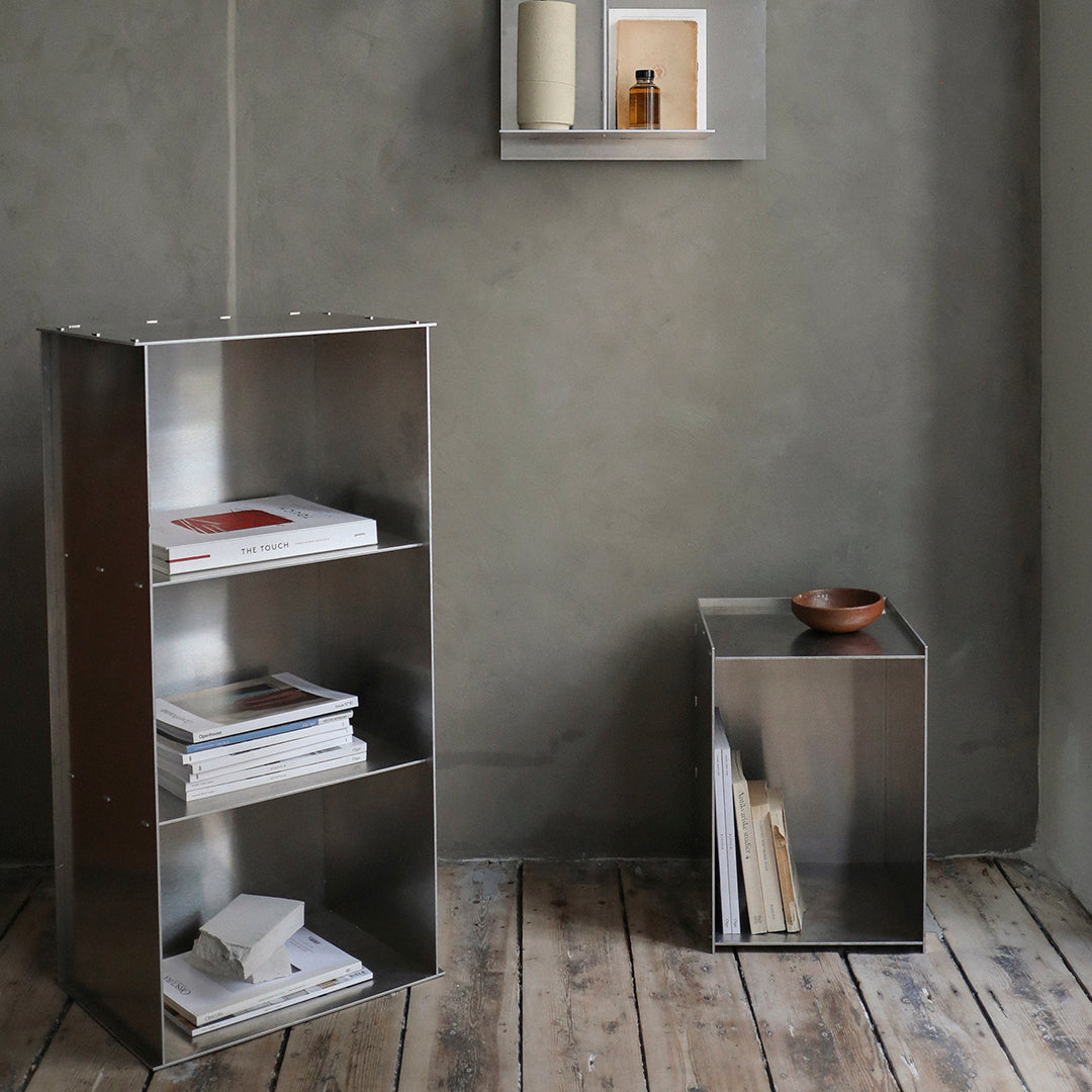 Metallic silver bookshelf and side table against a gray wall, showcasing sleek minimalist design with a wooden floor.