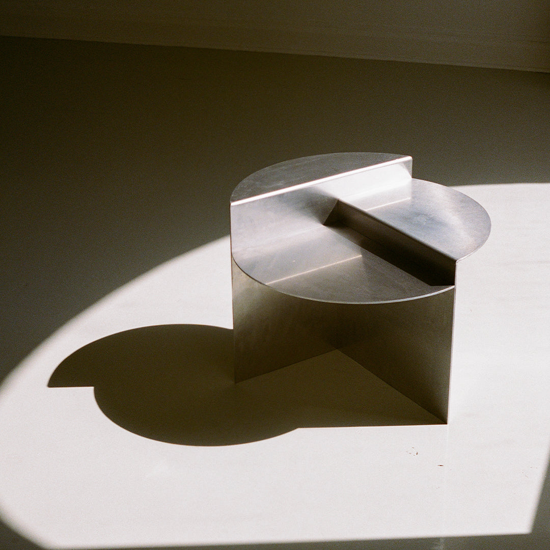 Sleek metallic stool with curved top and angular accents, casting soft shadows on a light surface.